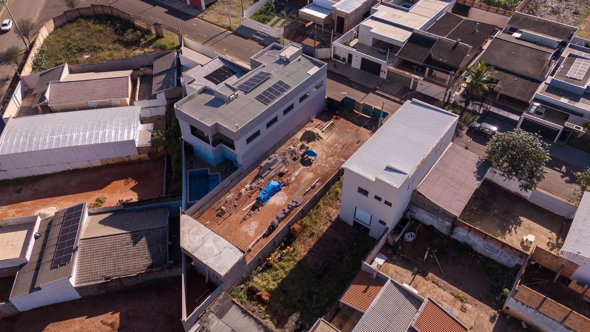 Vista aérea de uma obra residencial em andamento, com piscina e placas solares, cercada por casas do bairro